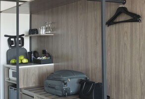 A compact hotel room with a wooden wall, a luggage rack, a small desk with a kettle, a coffee maker, and a few apples.