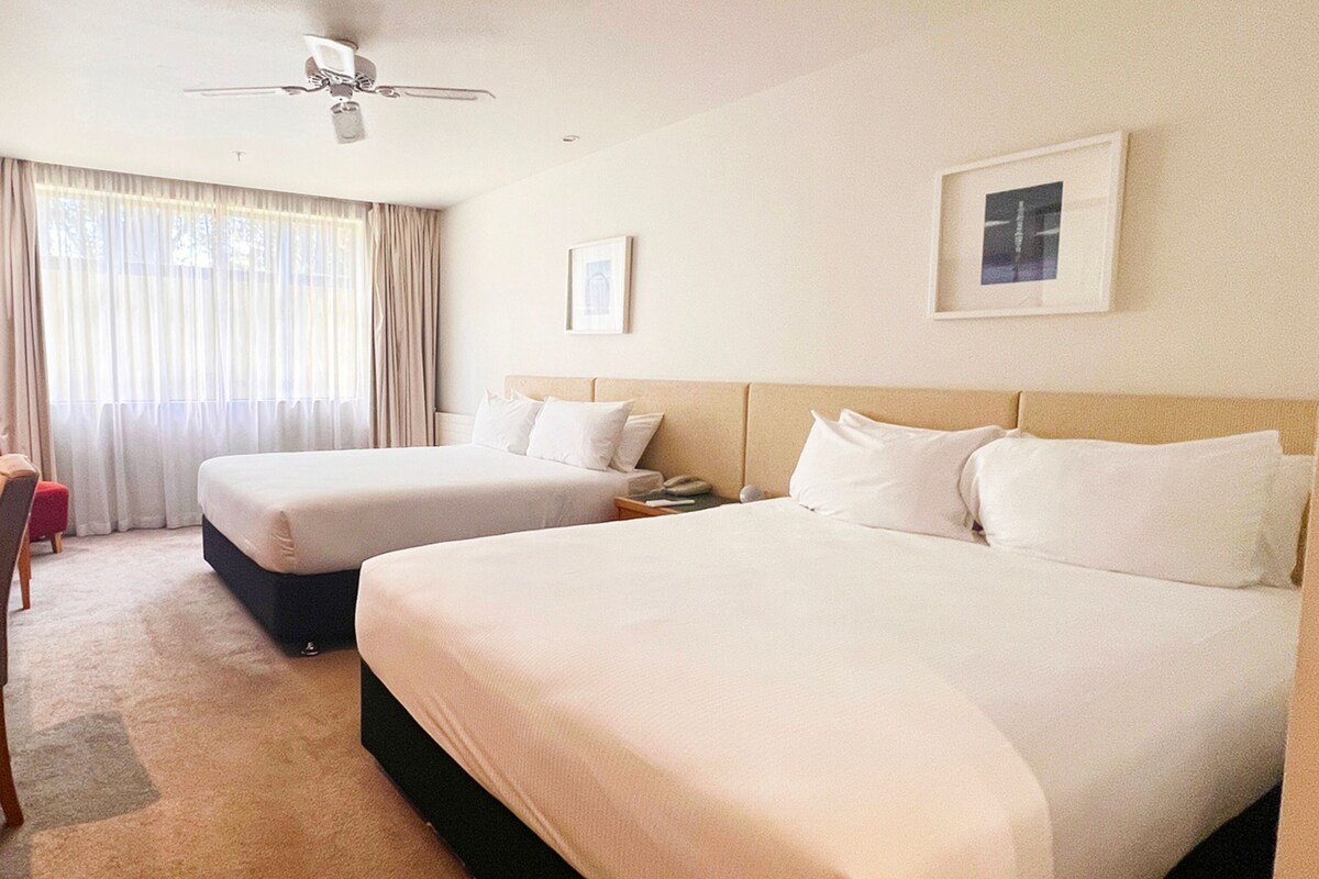 A hotel room with two beds, a ceiling fan, a window with curtains, and framed pictures on the wall.