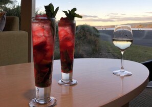 Twee hoge glazen met ijs en fruit op een houten tafel, en een wijnglas op een ronde tafel met uitzicht op water en heuvels.