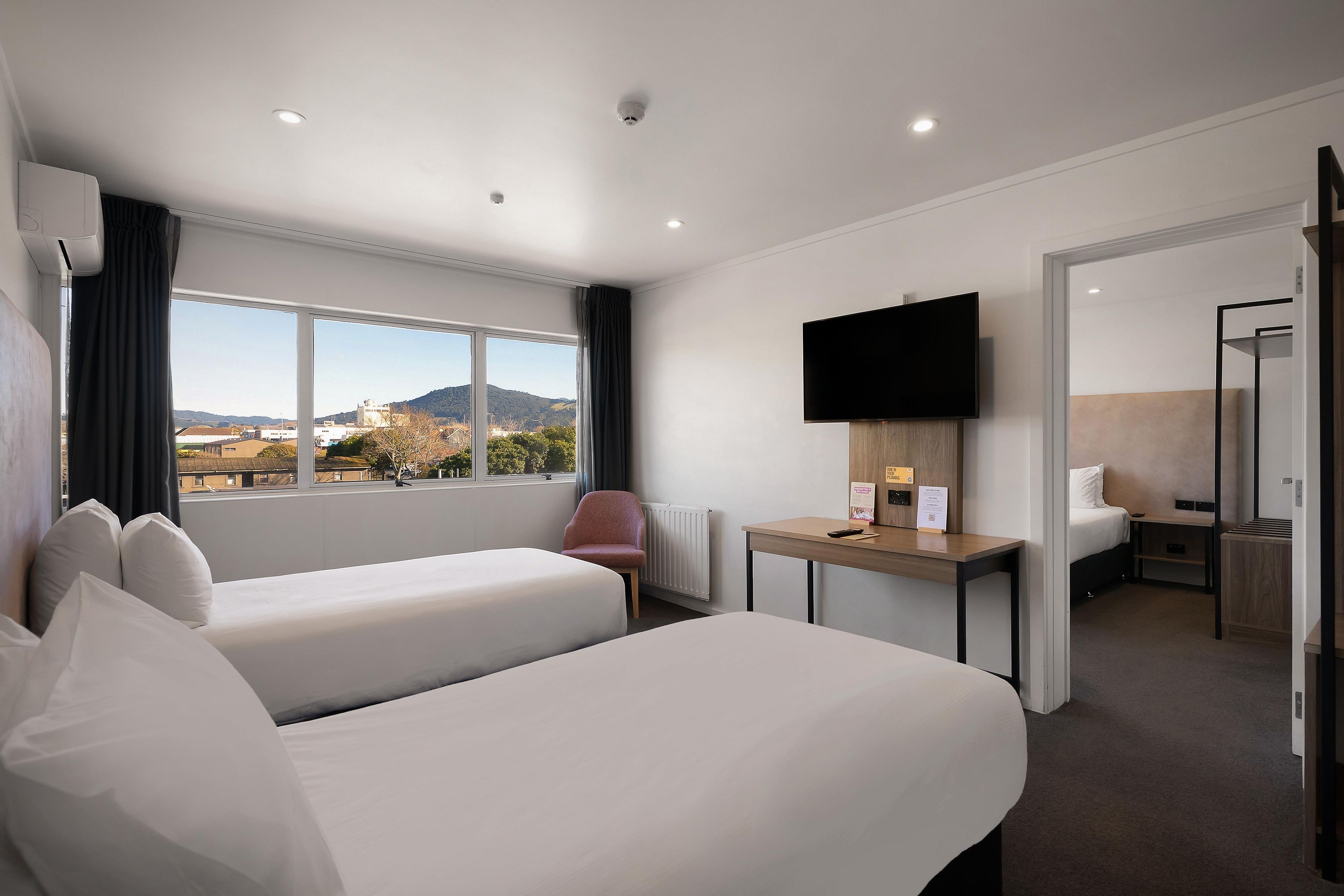 A hotel room with two beds, a desk, a TV, and a view of the outdoors.