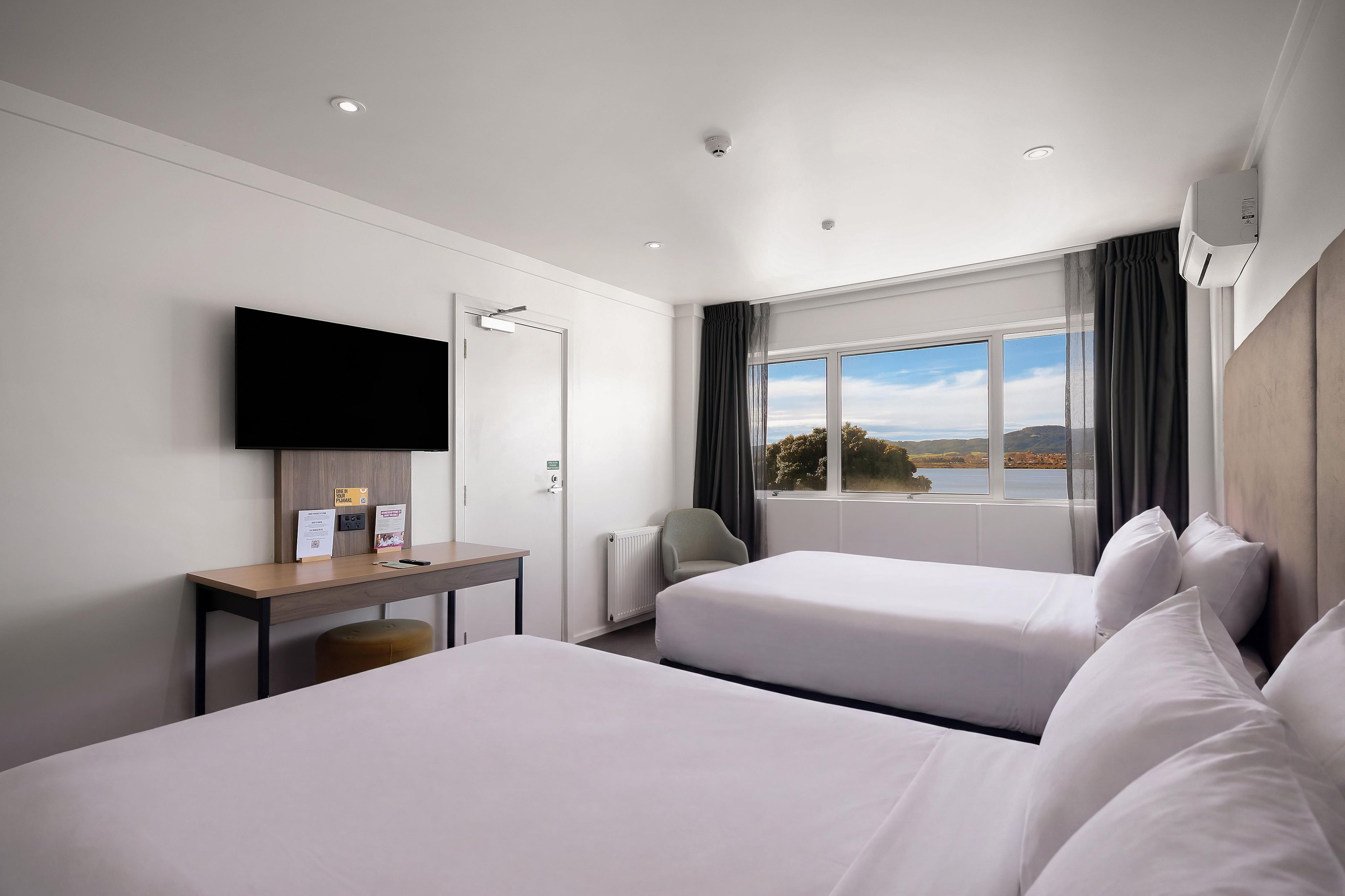 A hotel room with two beds, a desk, a flat-screen TV, a window with a view, and a thermostat.