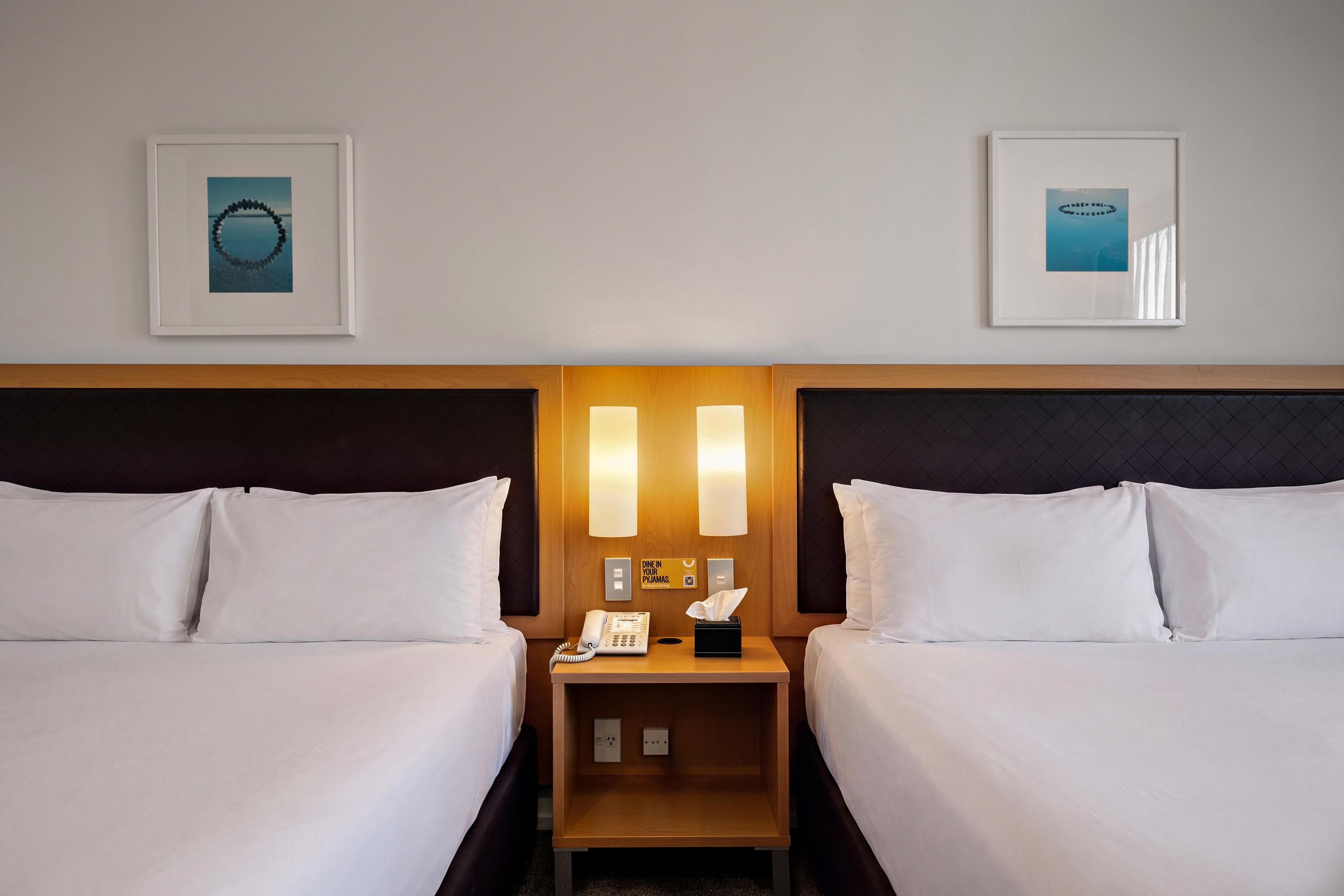 A hotel room with two beds, a nightstand, a phone, a tissue box, and two framed pictures on the wall.