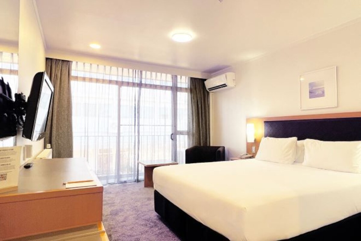 Habitación de hotel con una cama grande, un escritorio con computadora, una silla y un ventanal con cortinas.