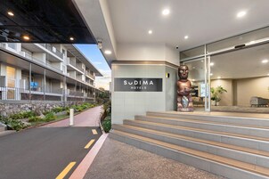 A hotel entrance with a sign for 'Sudima Hotels', a large statue, and a staircase leading to the lobby.