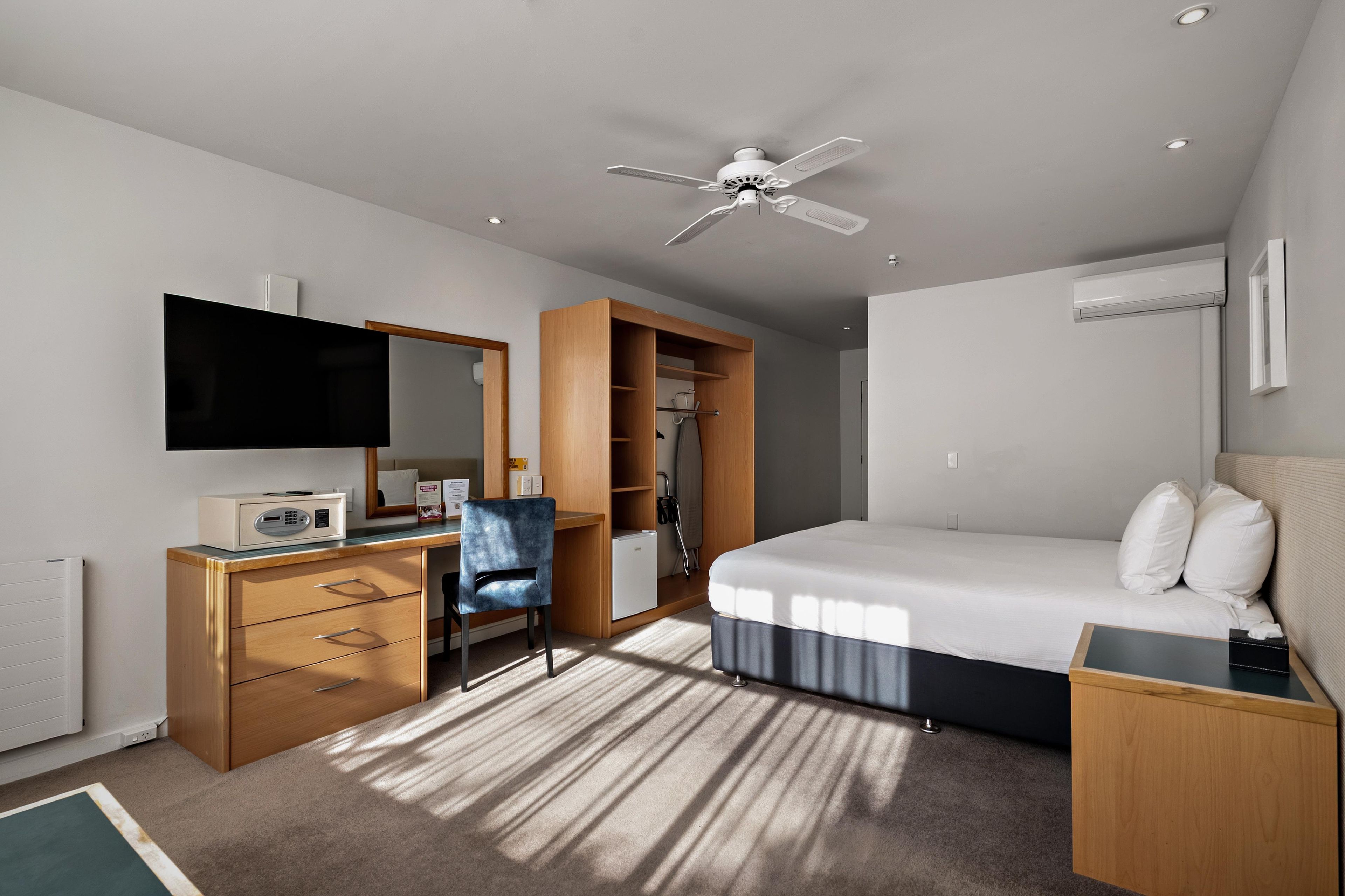 A modern hotel room with a flat-screen TV, a desk with a computer, a chair, a bed with white linens, a ceiling fan, and a built-in wardrobe.