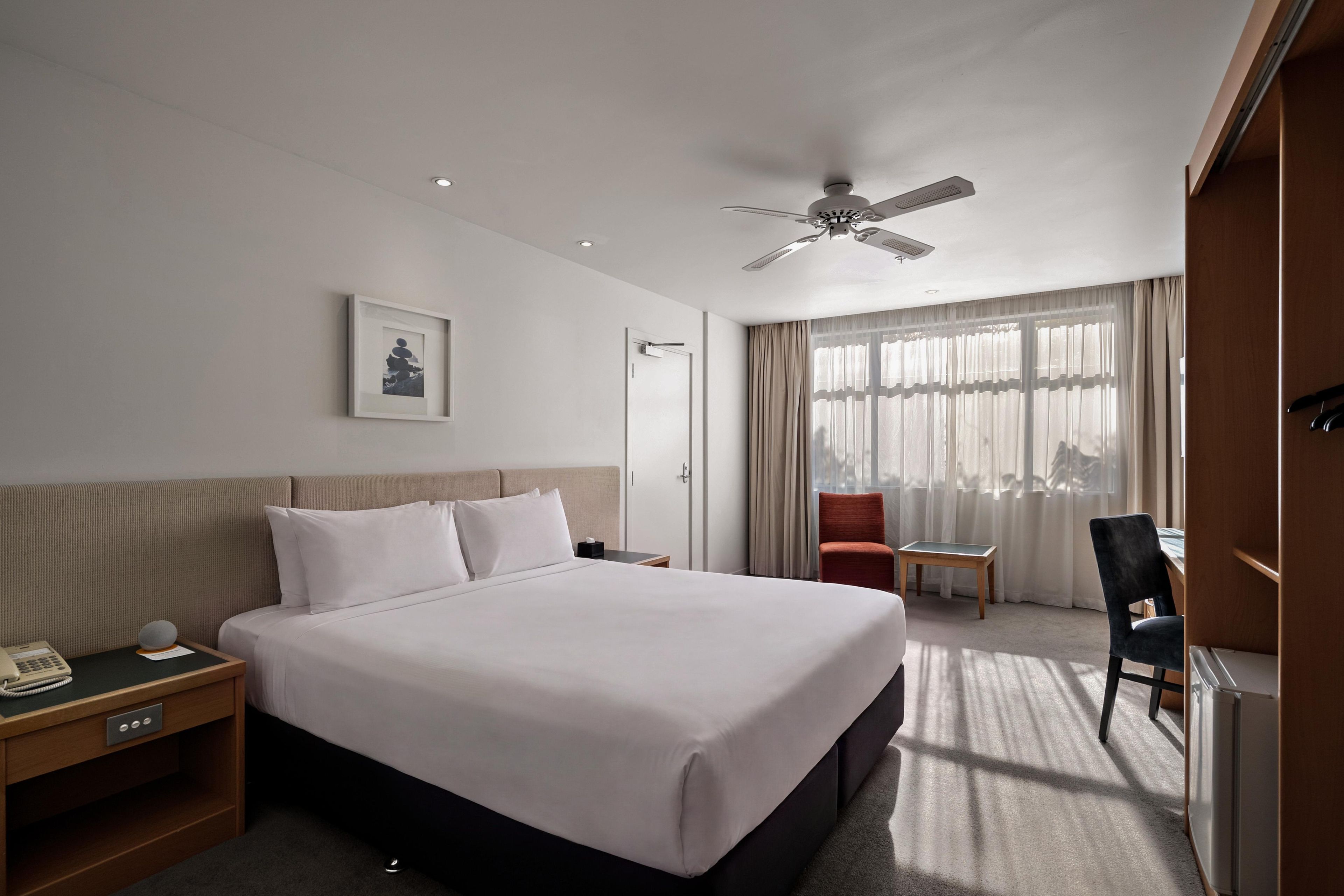 Ein Hotelzimmer mit Bett, Nachttisch, Stuhl, Schreibtisch und Deckenventilator.
