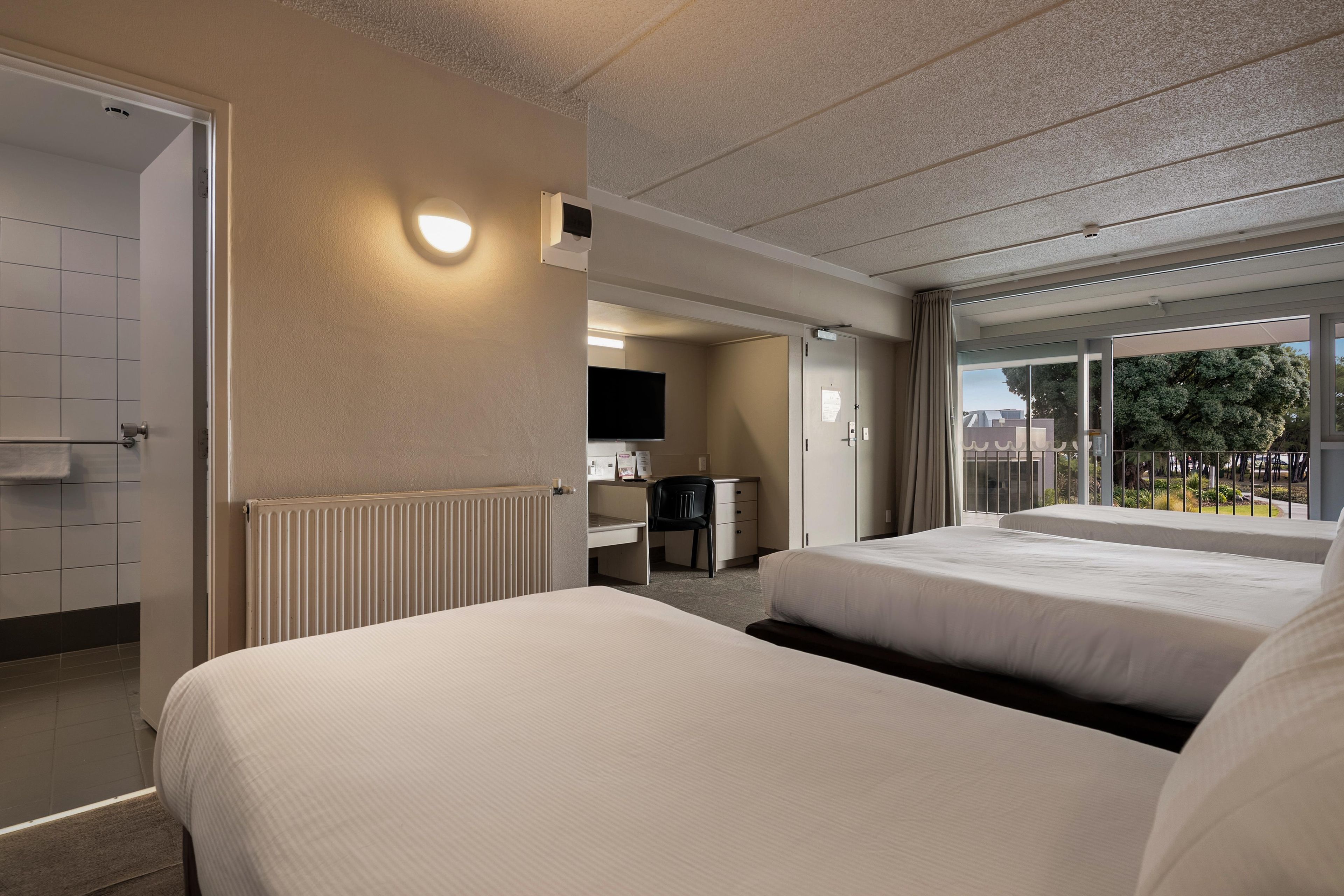 A hotel room with two beds, a desk, a television, and a balcony with a view.