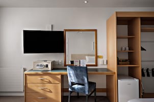 Quarto de hotel com escrivaninha de madeira, uma cadeira, televisão, espelho, guarda-roupa e um pequeno frigorífico.