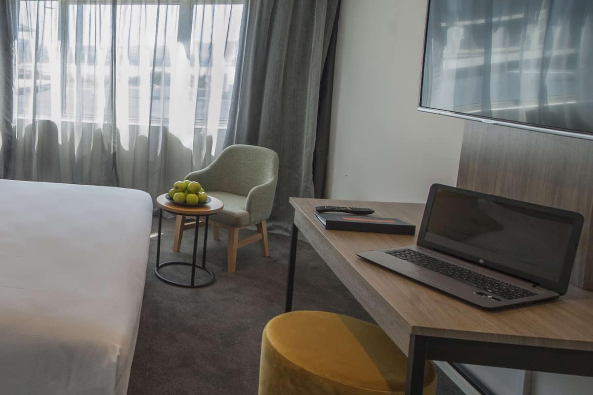 A hotel room with a bed, a desk with a laptop, a chair, a small table with fruit, and a large window with curtains.