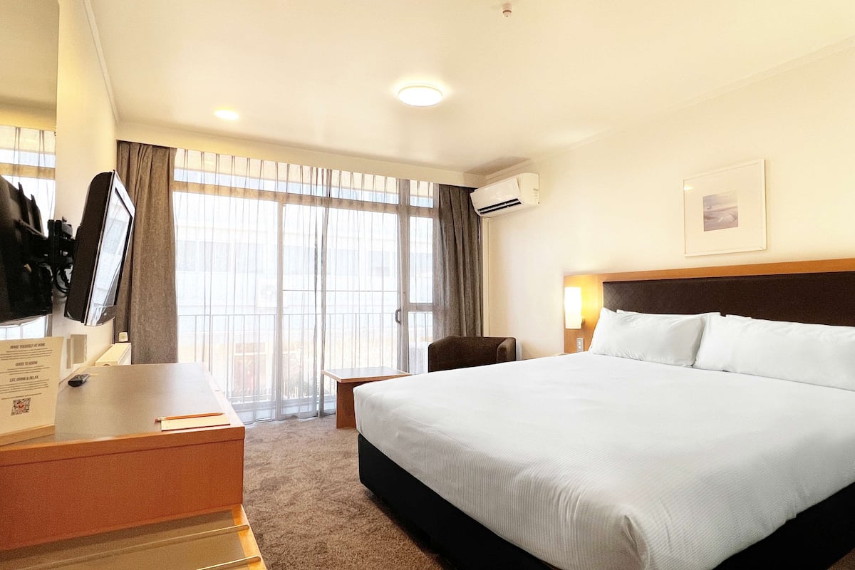 A hotel room with a large bed, a desk with a computer, a chair, and a window with curtains.