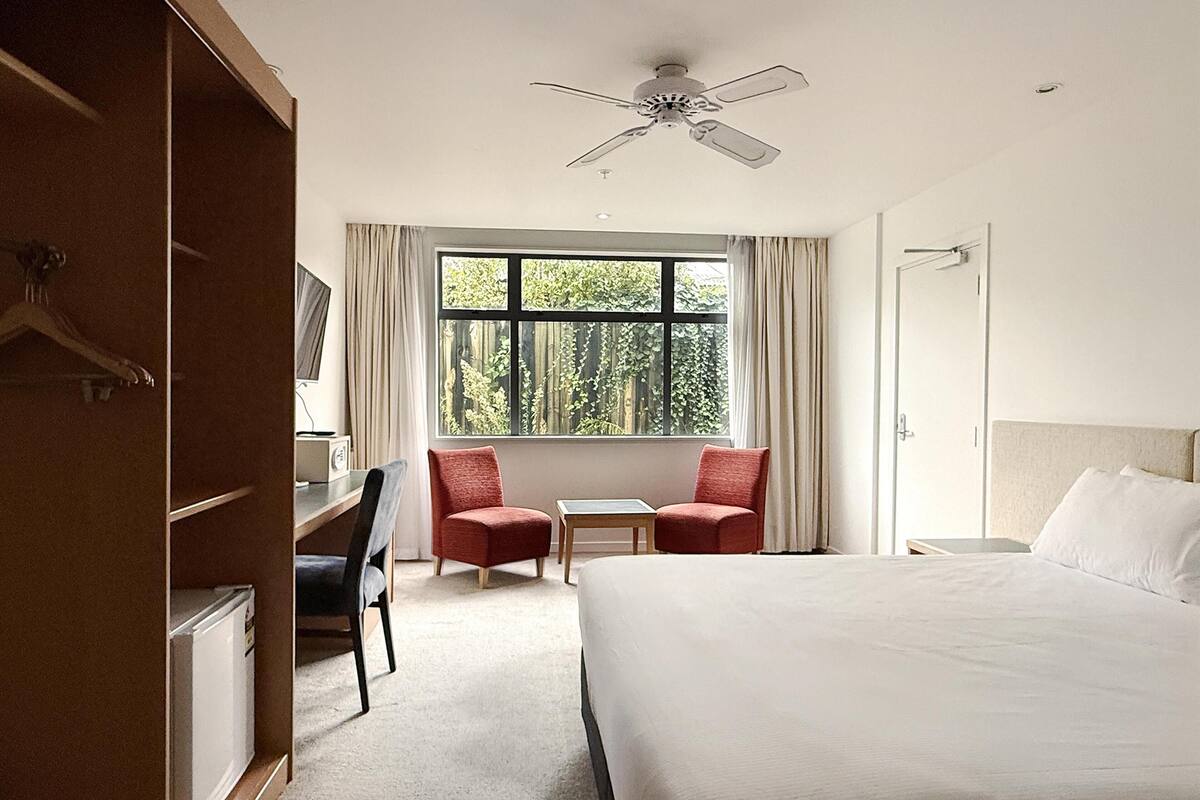 A hotel room with a bed, two red chairs, a small table, a desk with a chair, a wardrobe, and a window with curtains.
