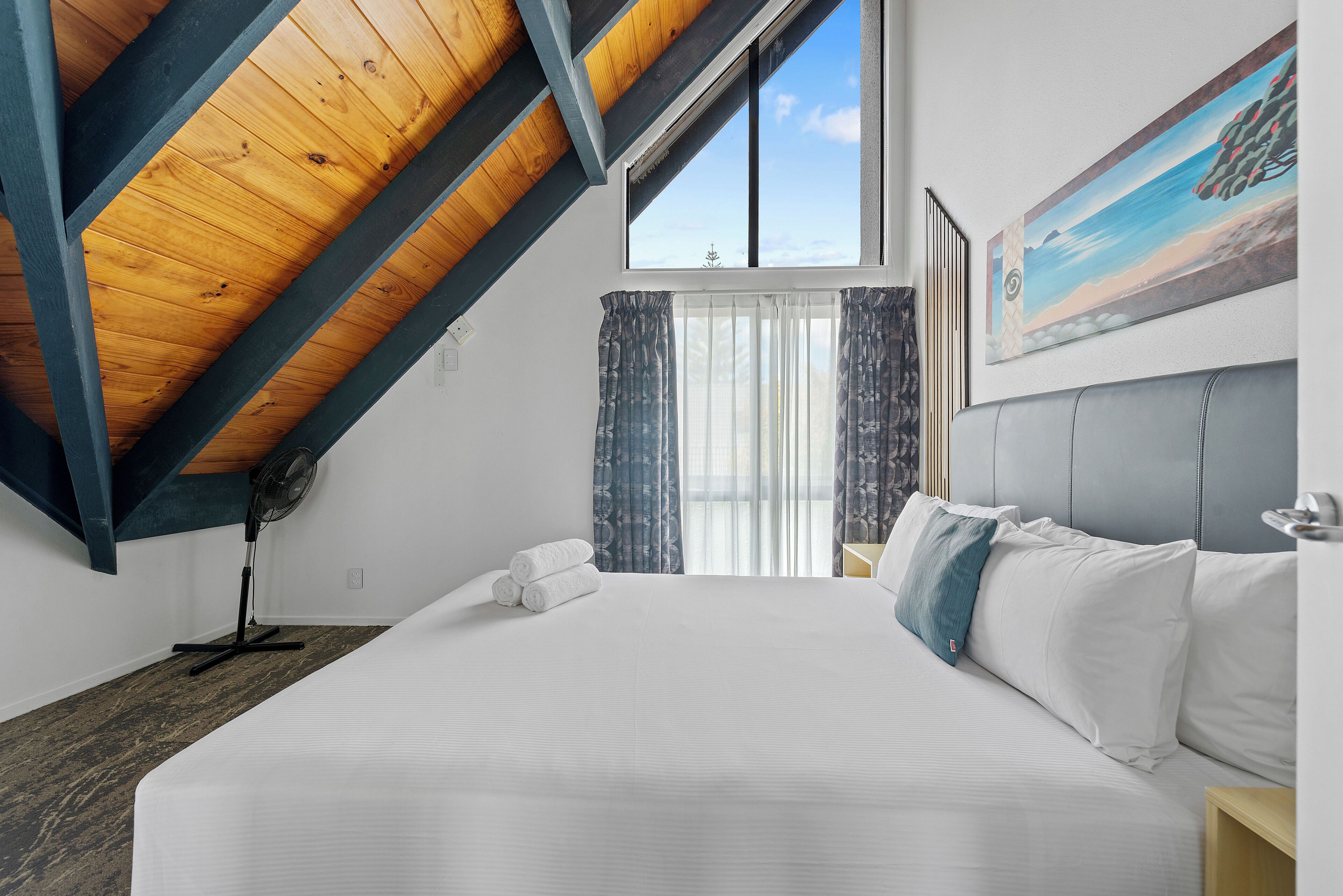A bedroom with a large bed, a ceiling fan, a window with curtains, and a painting on the wall.