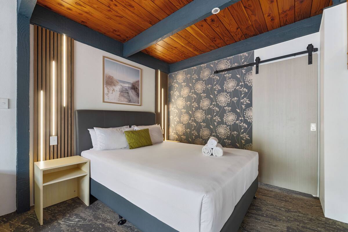 A neatly made bed with white linens and a green pillow, a wooden nightstand, a wall with floral wallpaper, and a ceiling with exposed wooden beams.