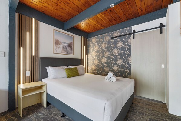 A neatly made bed with white linens and a green pillow, a wooden nightstand, a wall with floral wallpaper, and a ceiling with exposed wooden beams. - Wylie Court (Rotorua)
