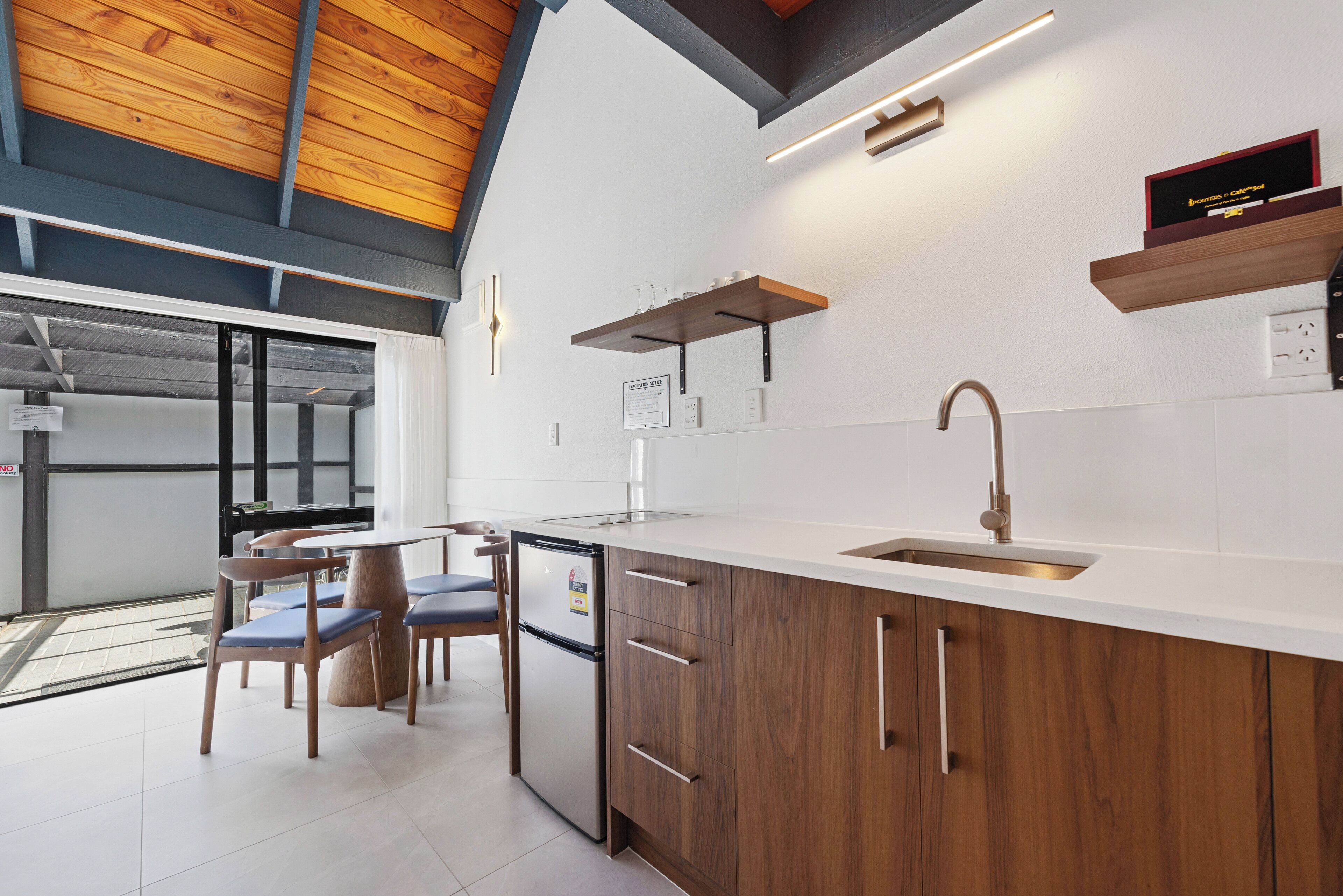 A modern kitchen with wooden cabinets, a stainless steel sink, and a built-in oven. There are chairs and a dining table in the adjacent area.