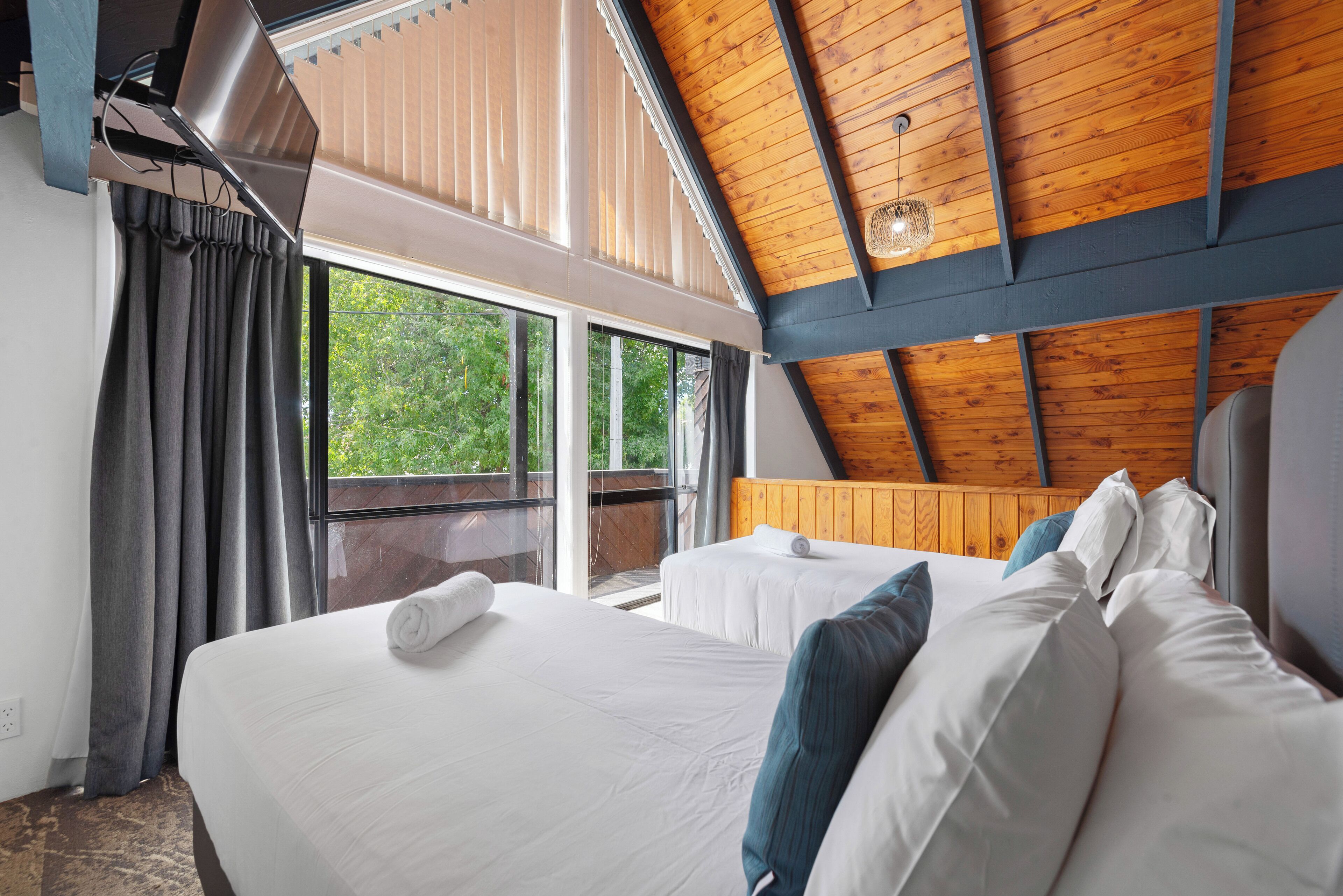 A modern hotel room with a sloped ceiling, large windows, and two beds with white linens and blue pillows.