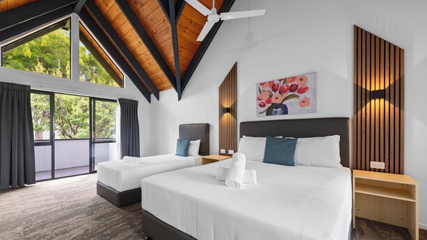 Quarto de hotel moderno com duas camas, janelas amplas, um ventilador de teto e um quadro na parede.