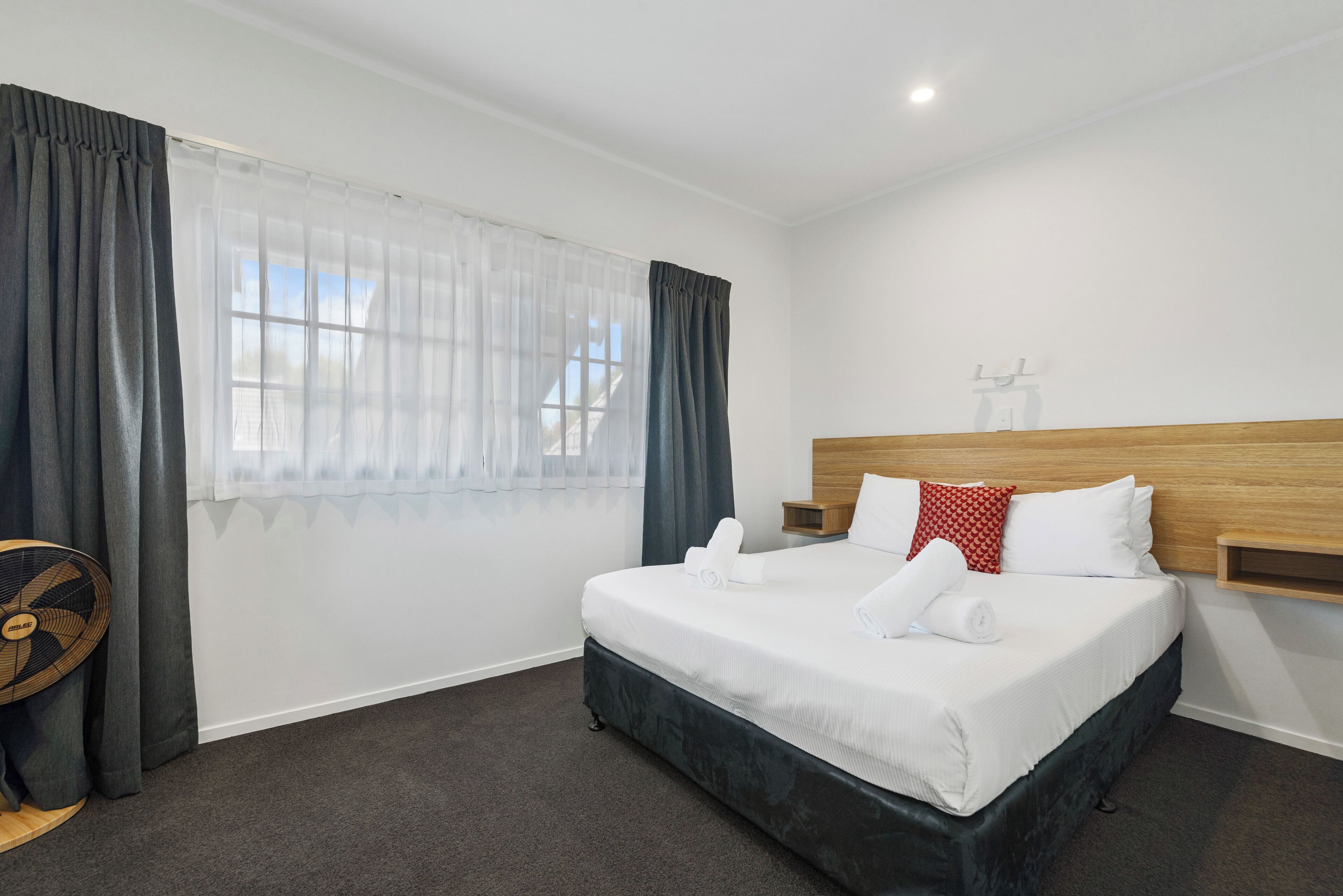 A neatly arranged hotel room with a bed, a bedside table, a fan, and a window with curtains.