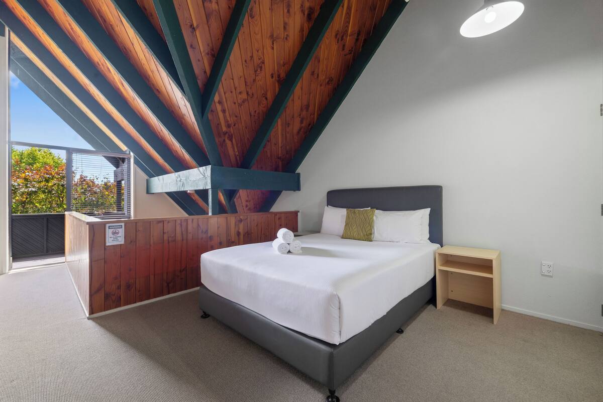 A bedroom with a large bed, a wooden headboard, a bedside table, and a view of the outdoors.