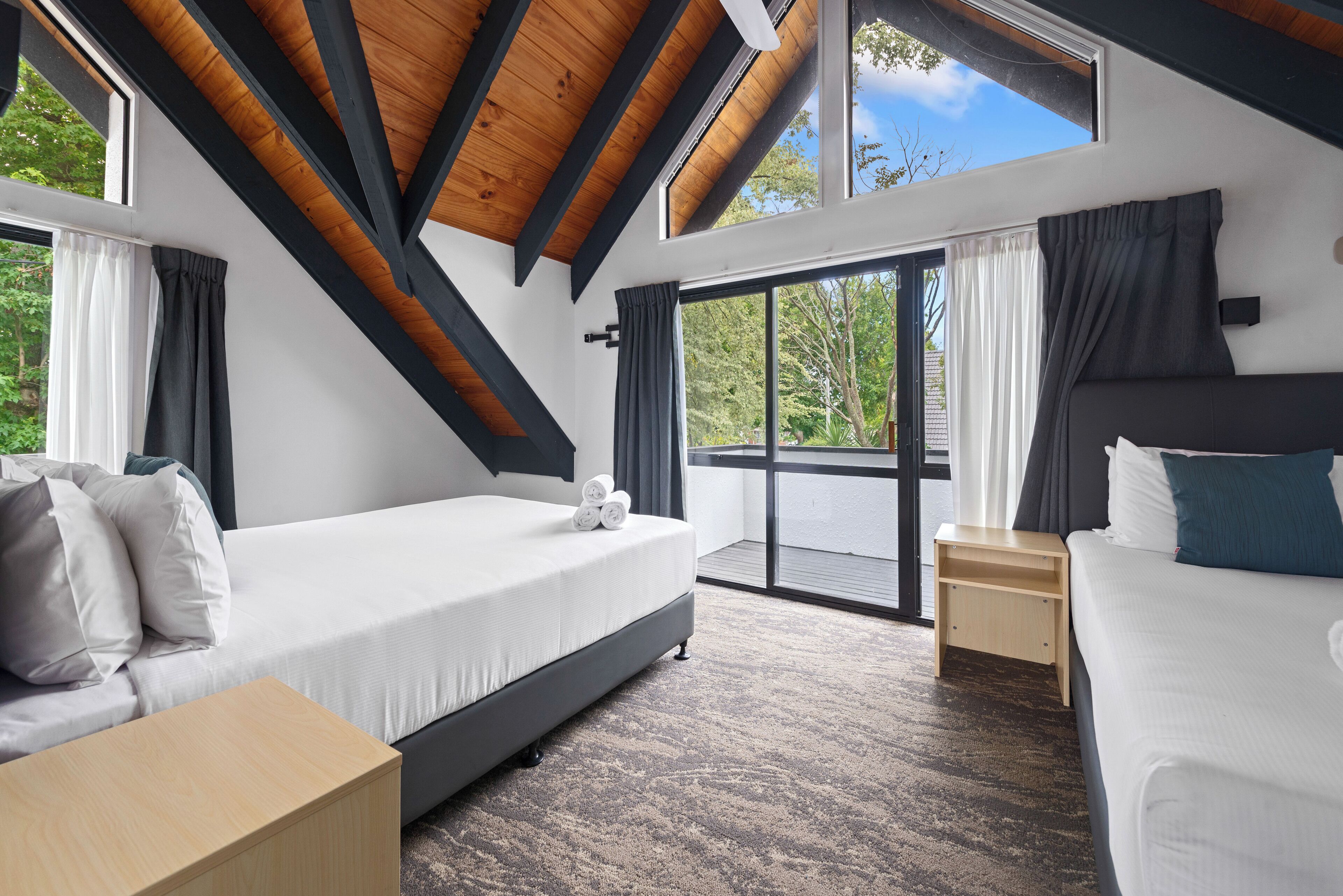 Une chambre moderne avec un grand lit, un lit plus petit, une table de chevet en bois et une vue sur l’extérieur grâce à de grandes fenêtres.