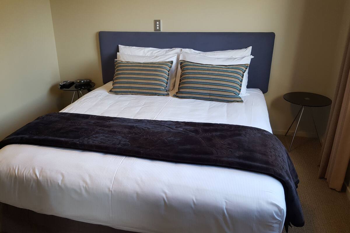 Un lit bien fait, avec du linge de lit blanc et des oreillers à rayures, une tête de lit bleu foncé et un plaid foncé.