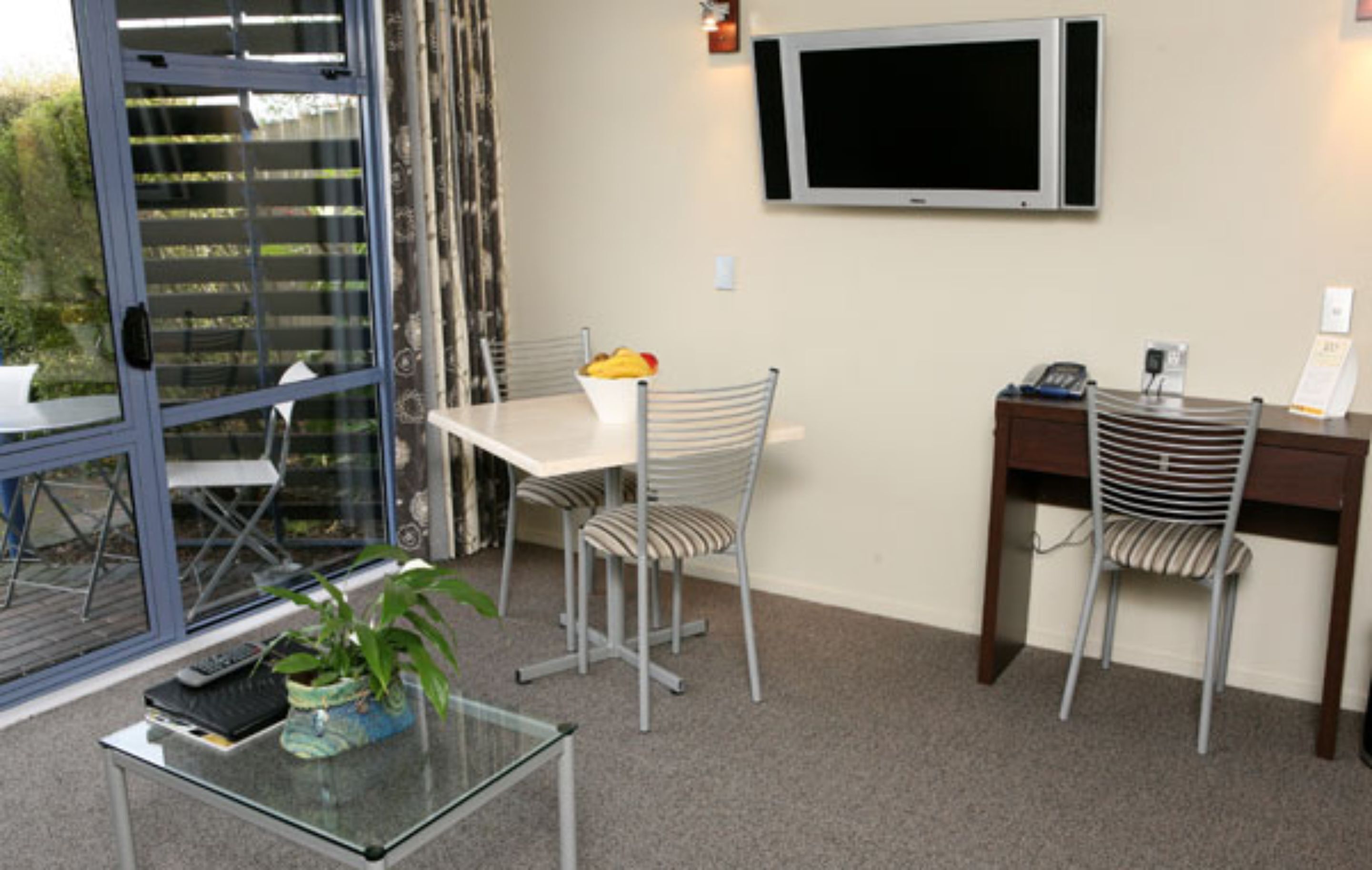 Una habitación con una mesa de vidrio, dos sillas, una pequeña planta en maceta, una mesa blanca, un televisor montado en la pared y un escritorio con una silla.