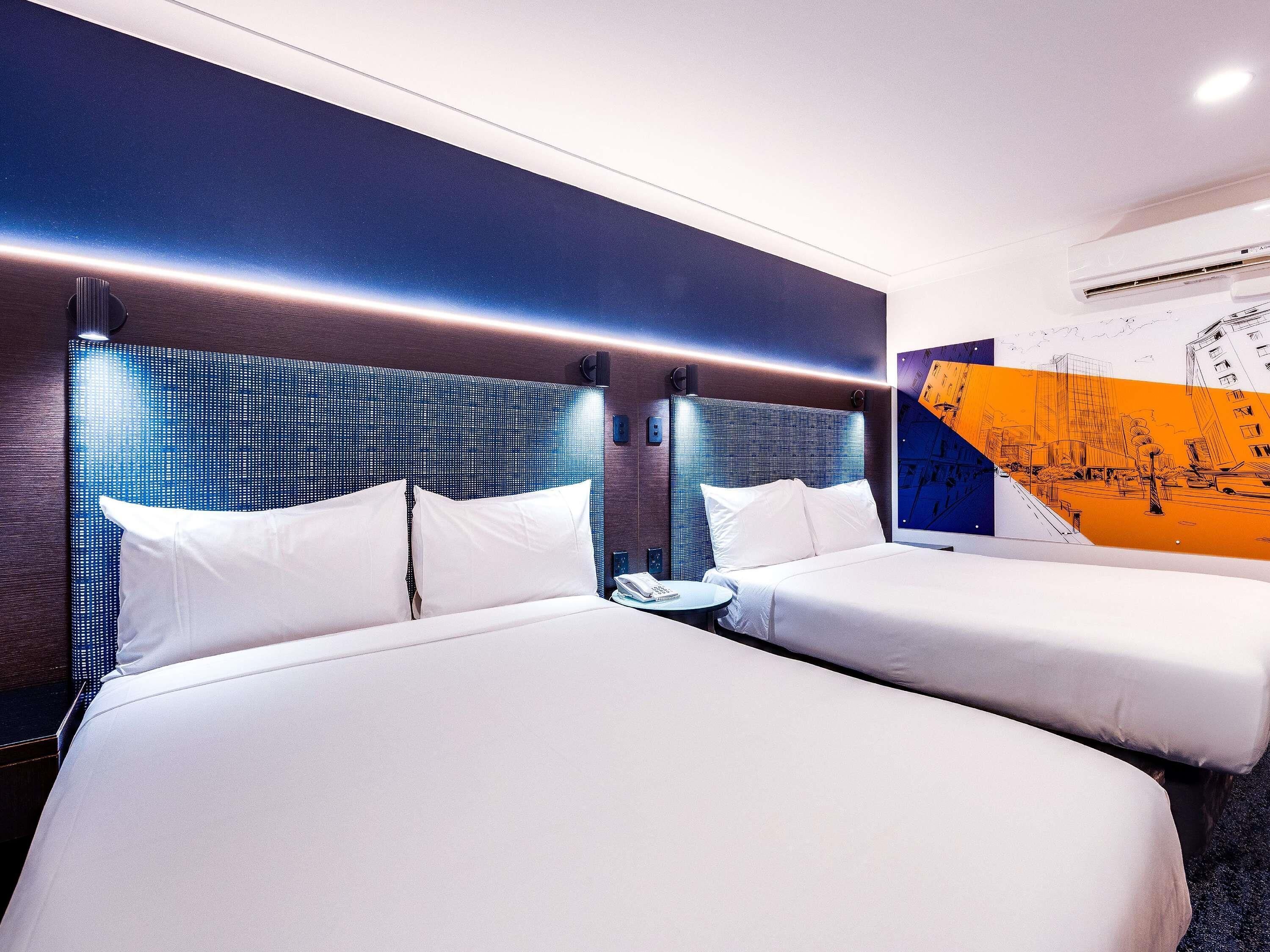 A hotel room with two beds, a wall-mounted air conditioner, and a cityscape mural.