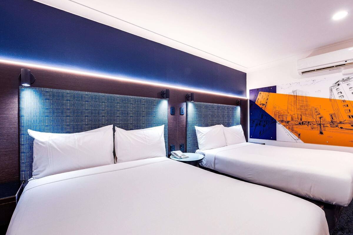 A hotel room with two beds, a wall-mounted air conditioner, and a cityscape mural.