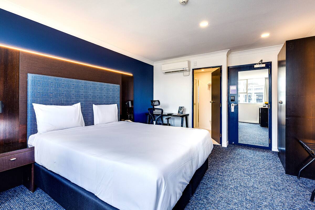 A hotel room with a large bed, a desk with a chair, a small table, and a door leading to another room.
