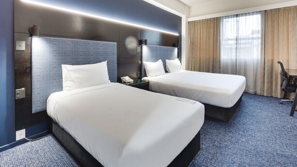 A hotel room with two beds, a desk, a chair, and a large window with curtains.