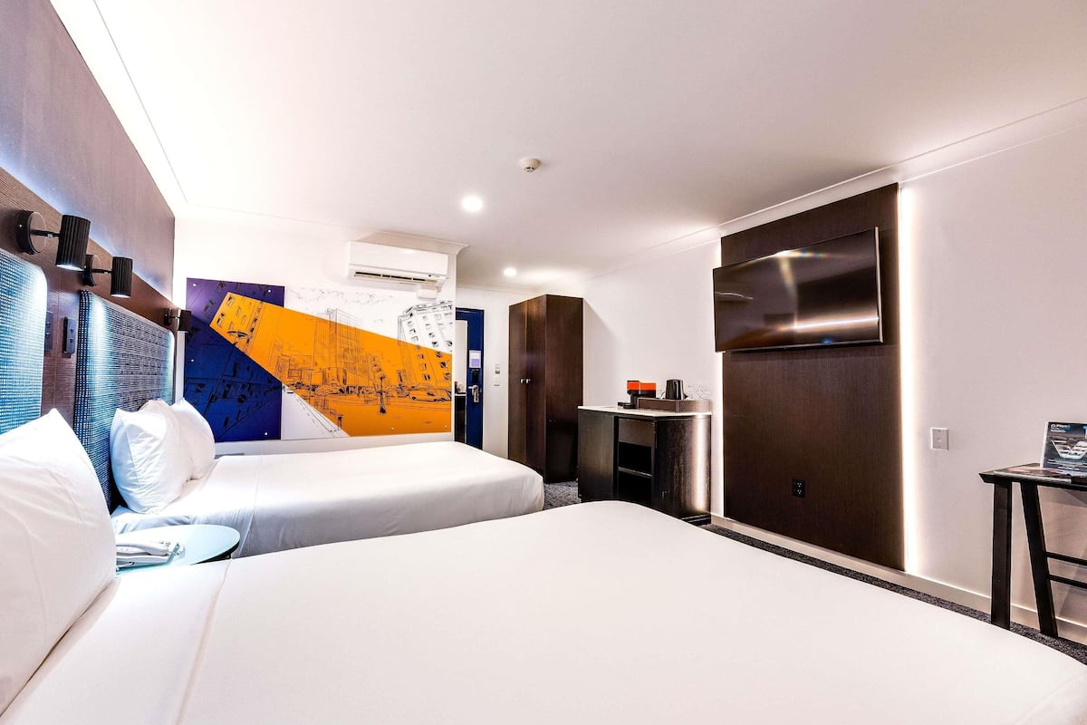 A modern hotel room with two beds, a desk, a flat-screen TV, and a large abstract wall mural.