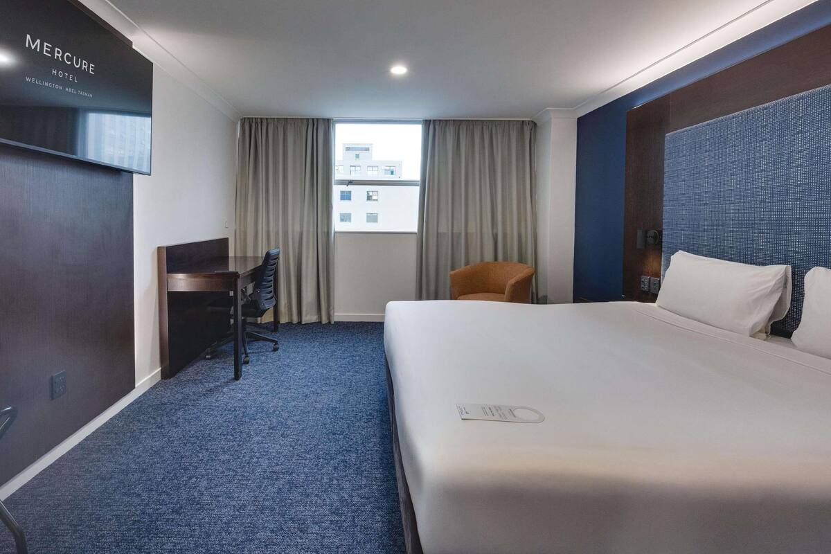 A hotel room with a bed, a desk, a chair, a TV, and a window with curtains.