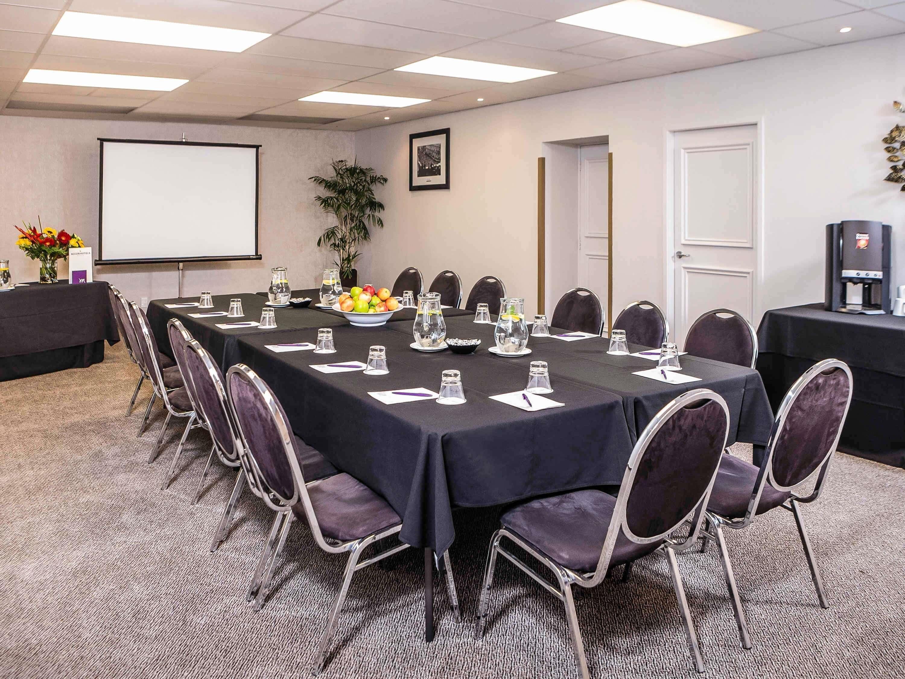 Un salón de conferencias con una larga mesa, sillas, una pantalla de proyector, una cafetera y una planta en maceta.