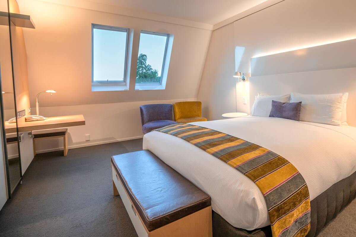 A hotel room with a bed, bedside table, desk, chair, and a window.