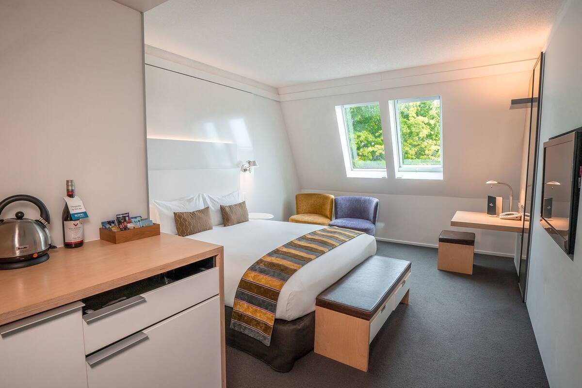A modern hotel room with a bed, bedside table, desk, chair, and a kettle.