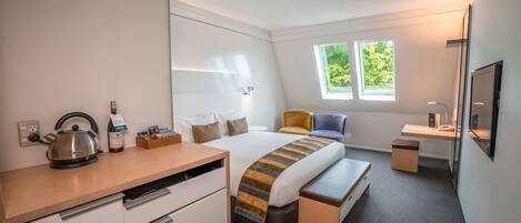 A modern hotel room with a bed, bedside table, desk, chair, and a kettle.