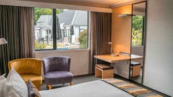 Une chambre d’hôtel avec un lit, deux chaises, un bureau, un miroir et une fenêtre donnant sur une maison et de la verdure.