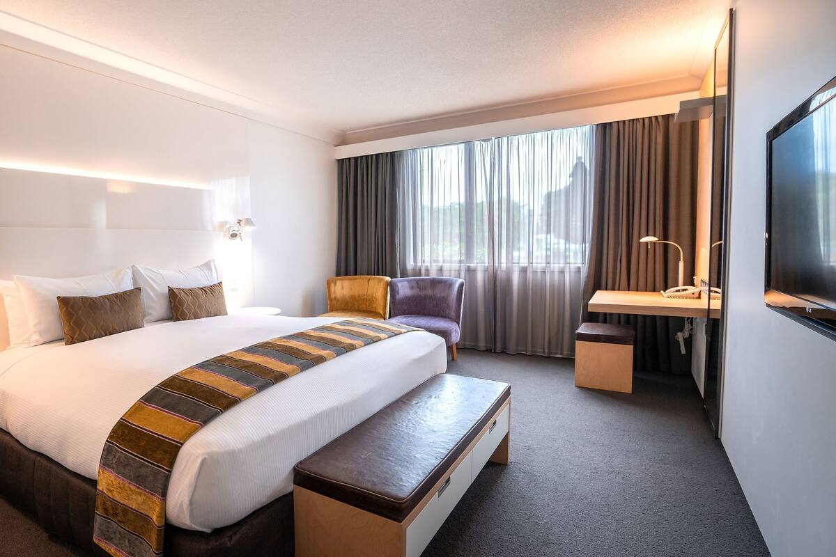 A hotel room with a large bed, bedside tables, a desk with a chair, a TV, and a window with curtains.