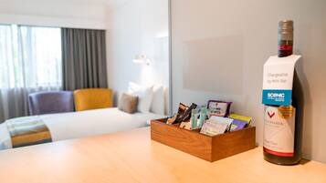 Una habitación de hotel con una cama, una mesita de madera con una botella de vino y una caja de bombones.