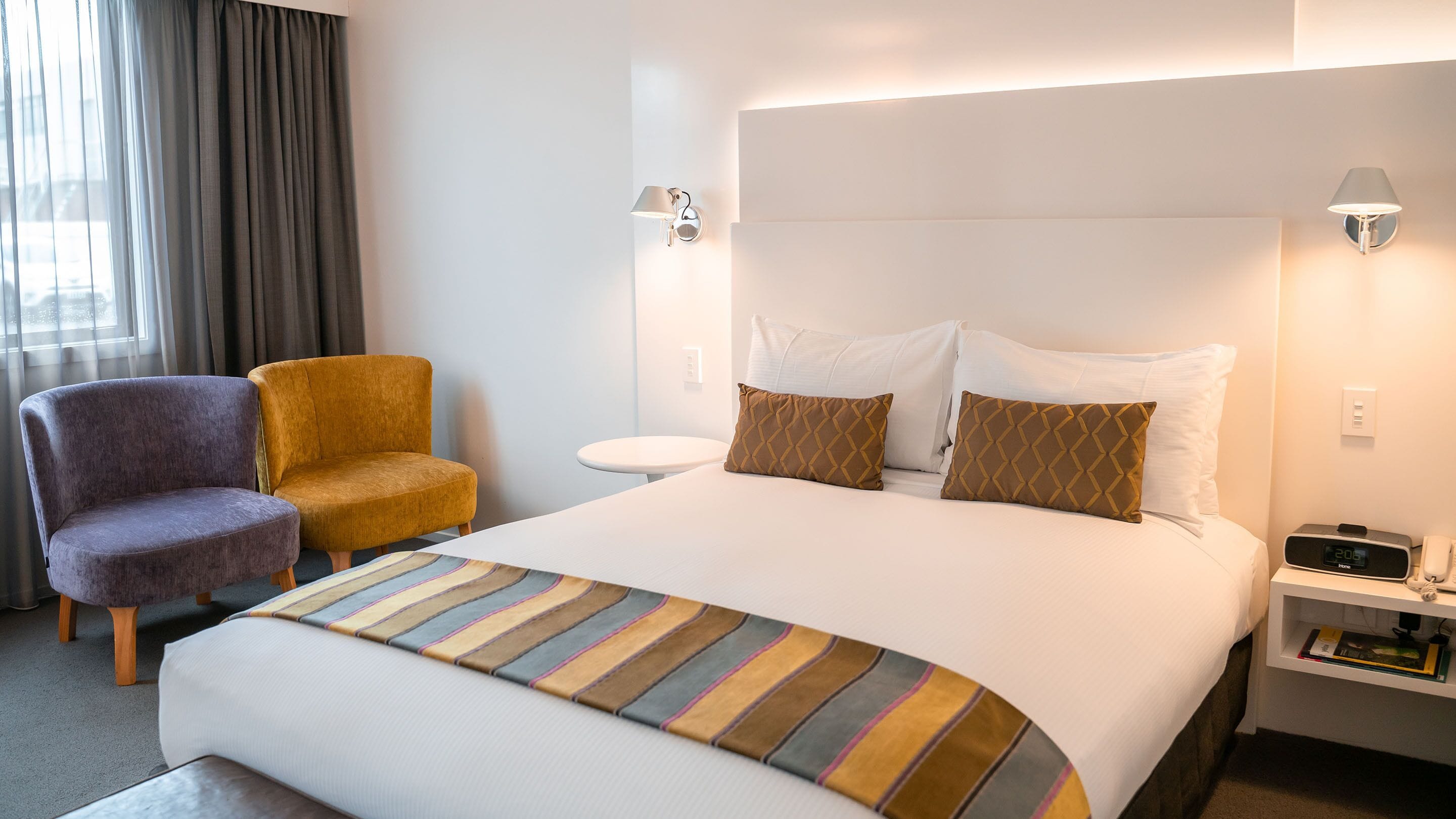 Een hotelkamer met een groot bed, twee stoelen, een nachtkastje, een lamp en een klok.