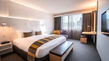 Une chambre d’hôtel comprenant un lit, une table de chevet, une lampe, une chaise, un bureau, une télévision et des rideaux.