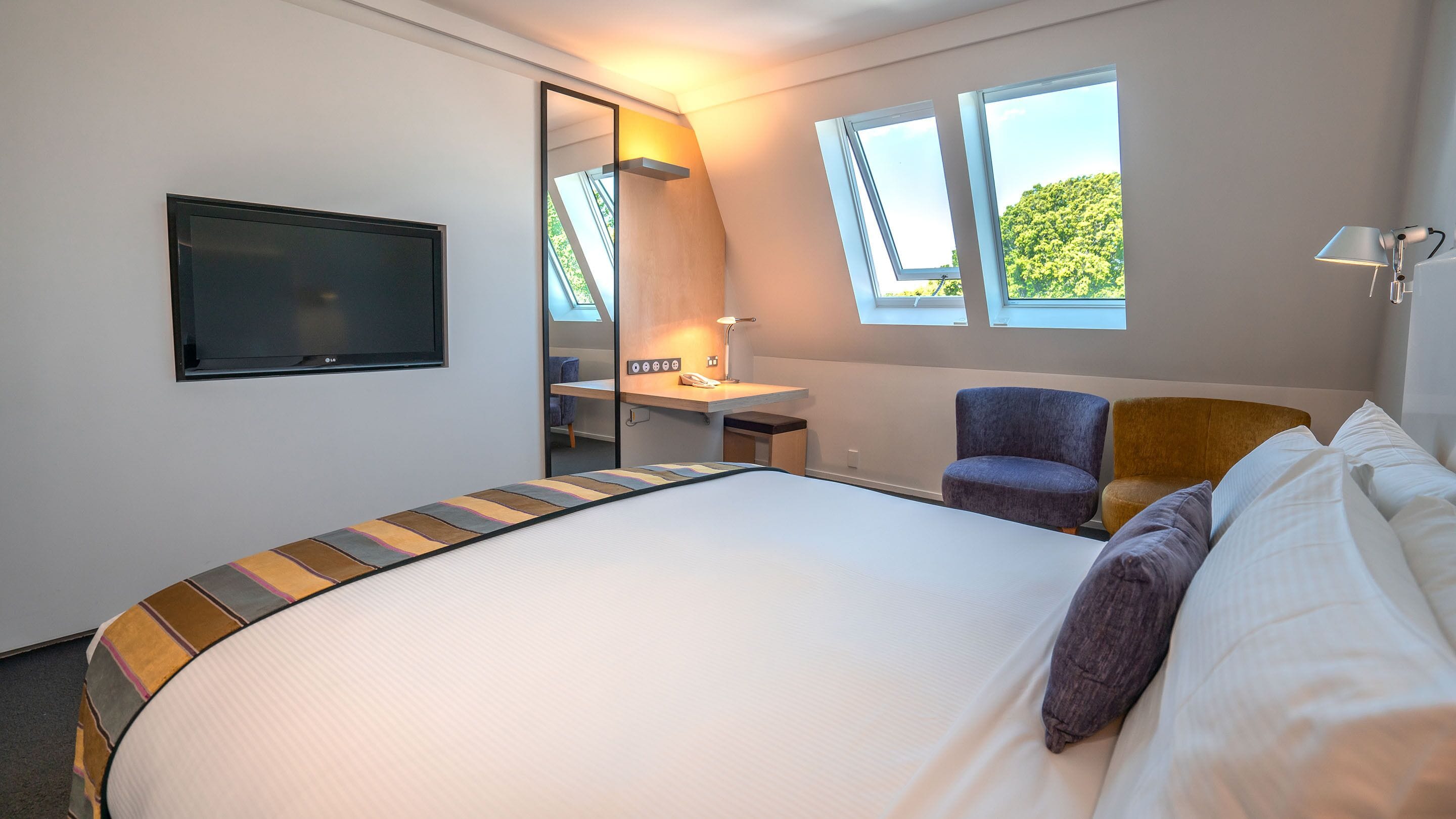 A hotel room with a large bed, a desk with a chair, a TV, and a window with a view of greenery.
