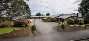 A motor inn with a driveway leading to a parking area, surrounded by greenery and a clear sky.