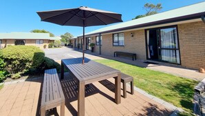 A patio area with a wooden table and an umbrella, surrounded by a brick building with large windows and a door. - ASURE Highpark Motor Inn (Greymouth)