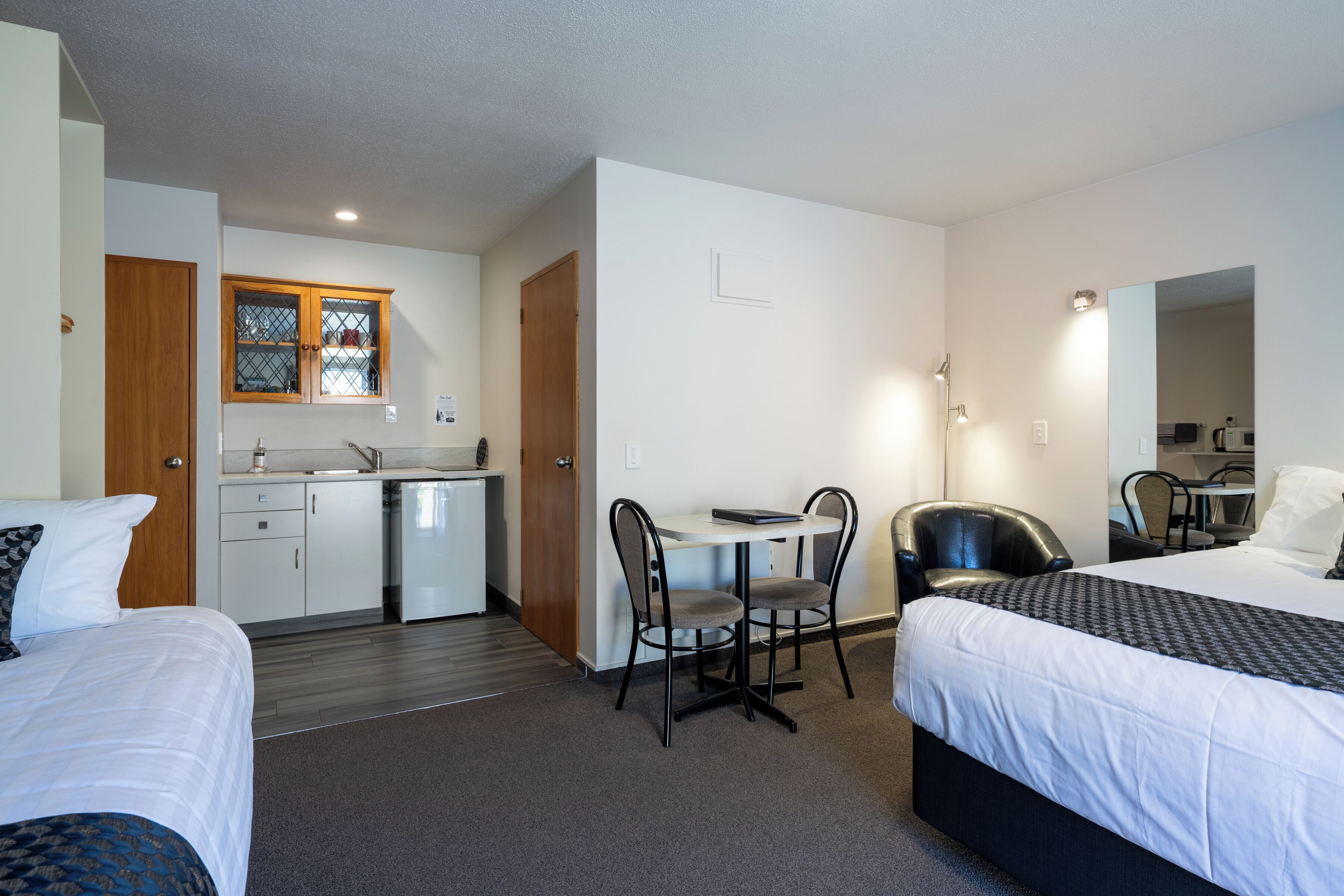 A hotel room with two beds, a desk with a chair, a small kitchenette, and a mirror.