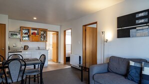 A modern living room with a grey sofa, a dining table with chairs, a kitchen area with cabinets, and a white radiator. - ASURE Highpark Motor Inn (Greymouth)