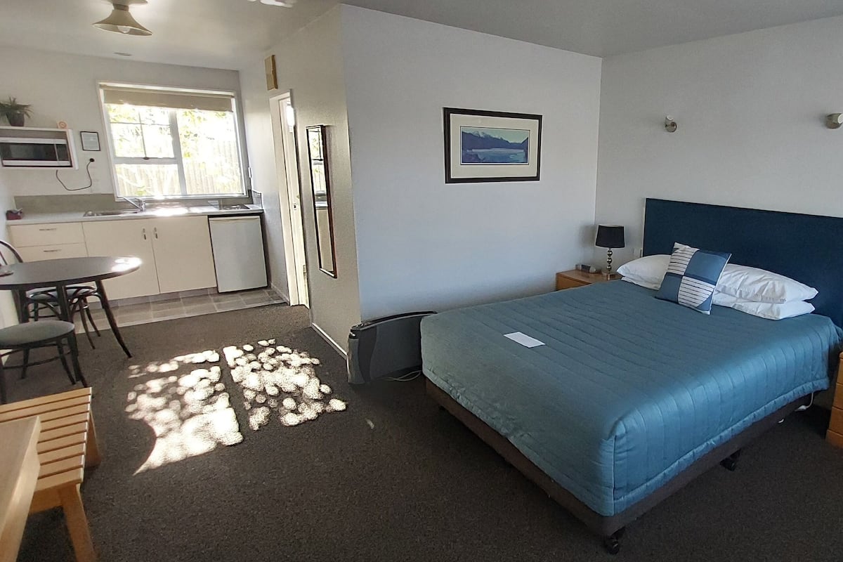 Quarto com cama, mesas de cabeceira, escrivaninha e uma cadeira. Há uma janela com vista para o exterior.