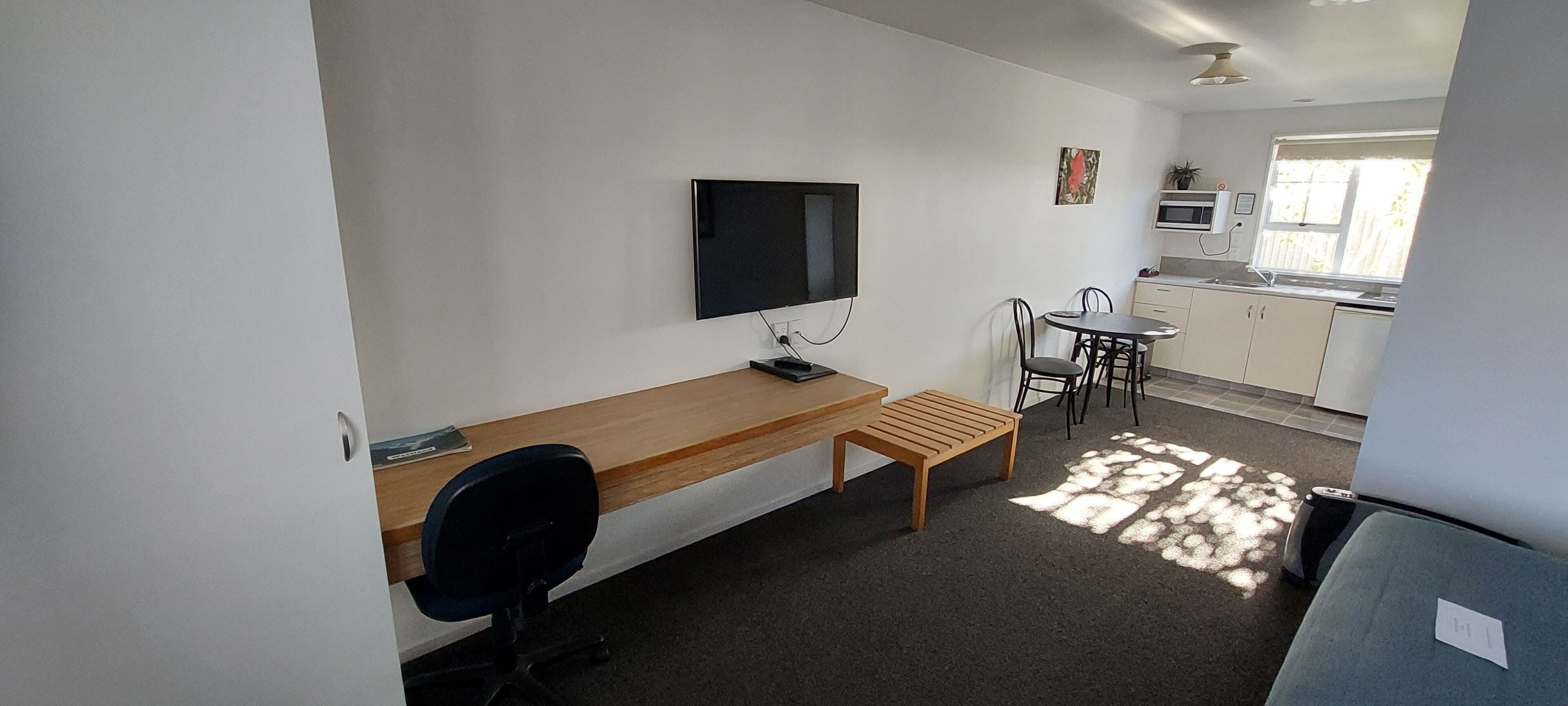 Ein gemütlicher Raum mit Schreibtisch aus Holz, einem Sessel, einem an der Wand montierten Fernseher, einer kleinen Küchenzeile mit Mikrowelle und einem Fenster mit Tageslicht.