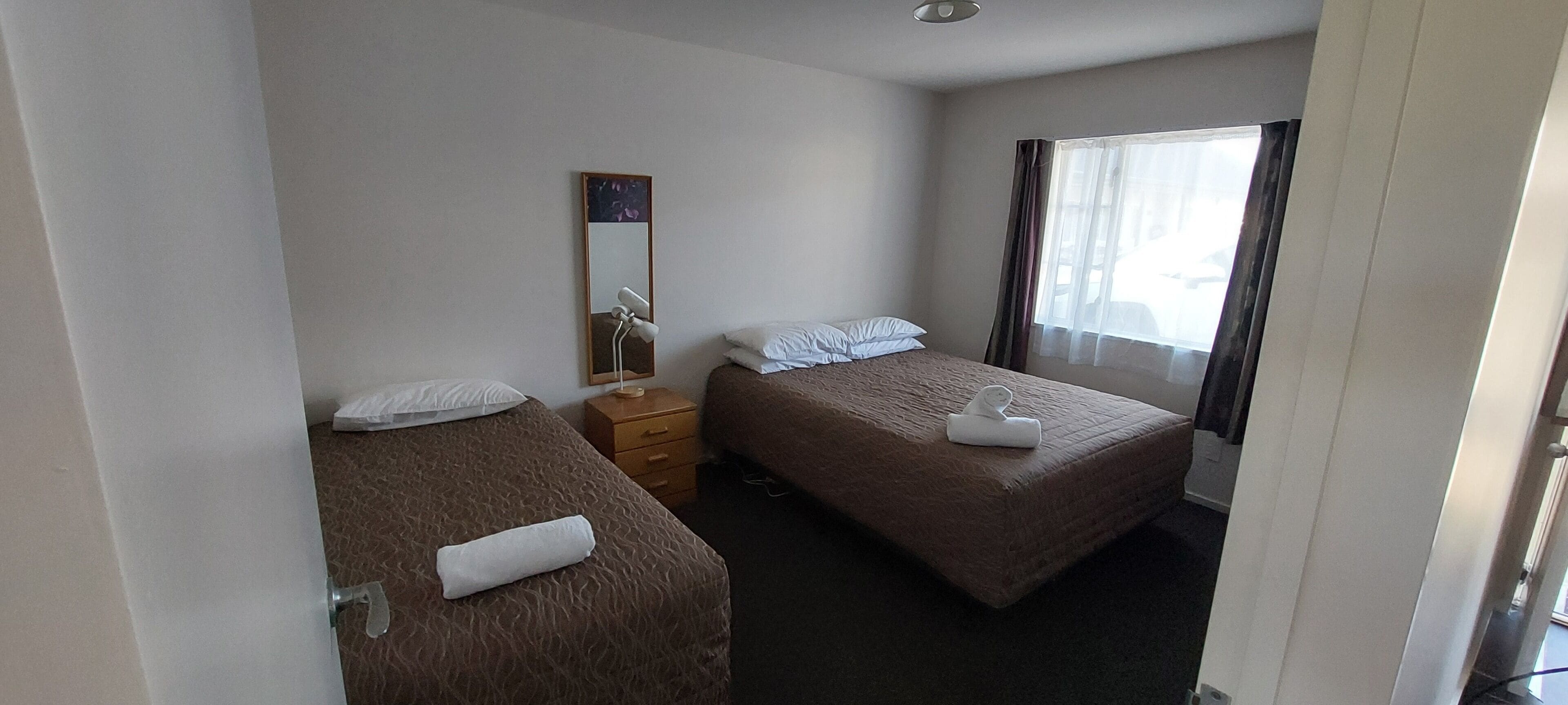 Een hotelkamer met twee eenpersoonsbedden, een nachtkastje met een lamp, een spiegel en een raam met gordijnen.