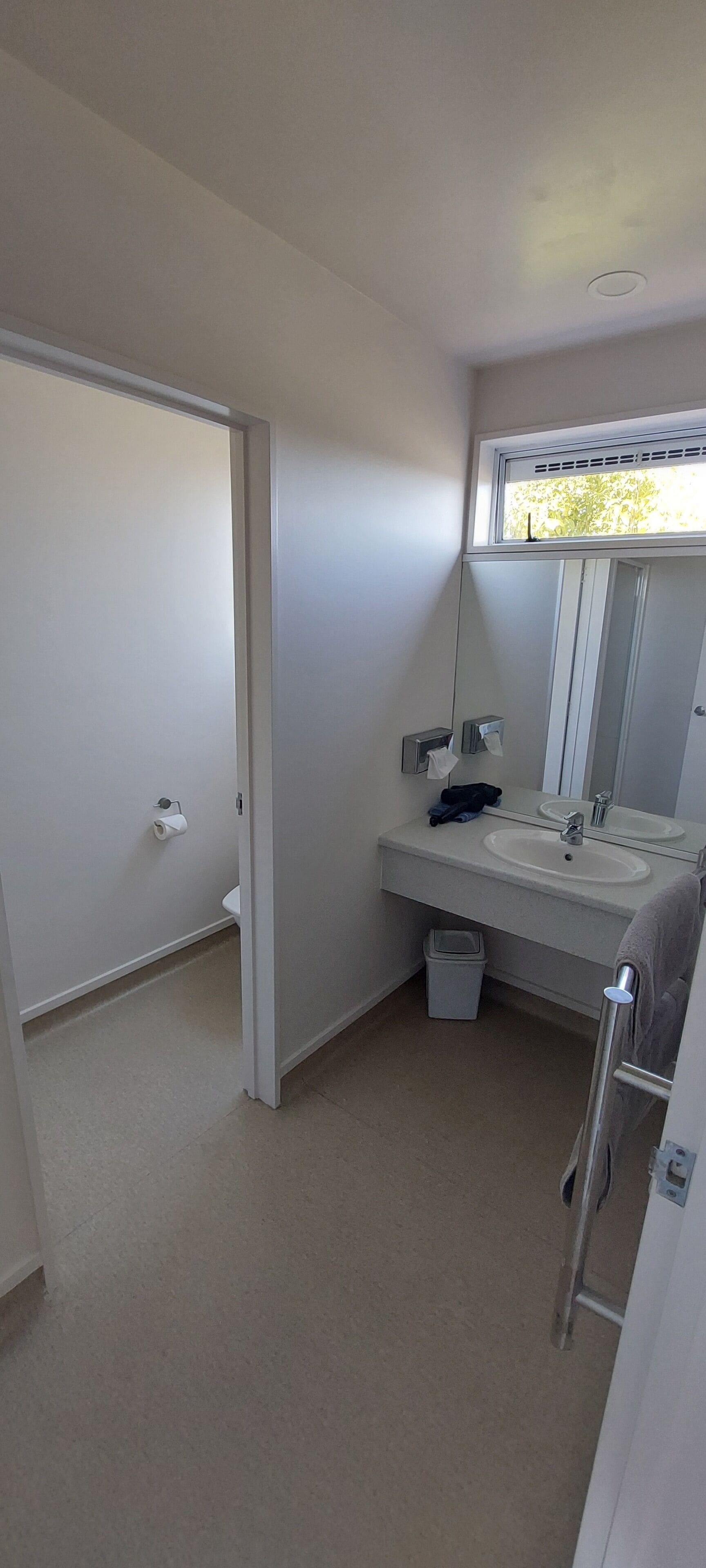 A bathroom with a toilet, sink, and mirror. There is a towel rack and a trash bin.
